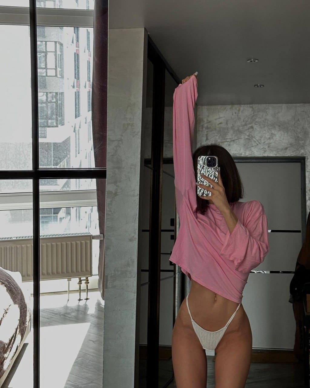 A woman stands by large windows in a cropped pink top and white bottoms, holding a phone for a mirror selfie.