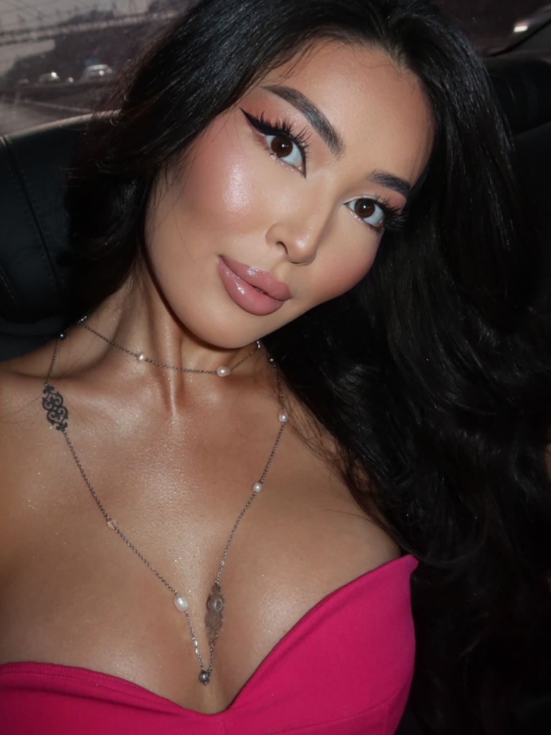 A close-up shows a woman in a pink strapless top and layered necklaces, seated in a dark room with a window behind her.
