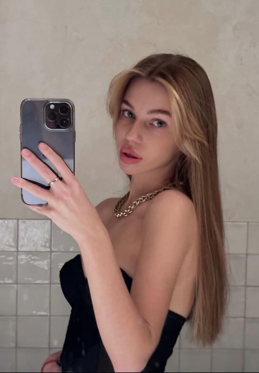 Taking a mirror selfie, she wears a black strapless top and gold chain against a tiled wall.