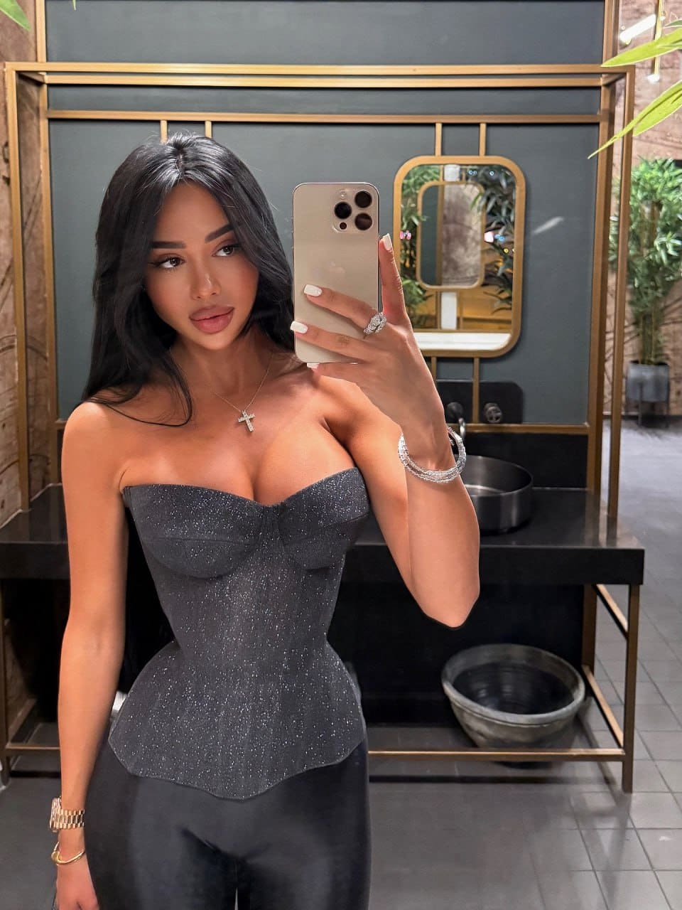 A woman takes a bathroom mirror selfie wearing a sparkly grey strapless top, dark pants, and a cross necklace.