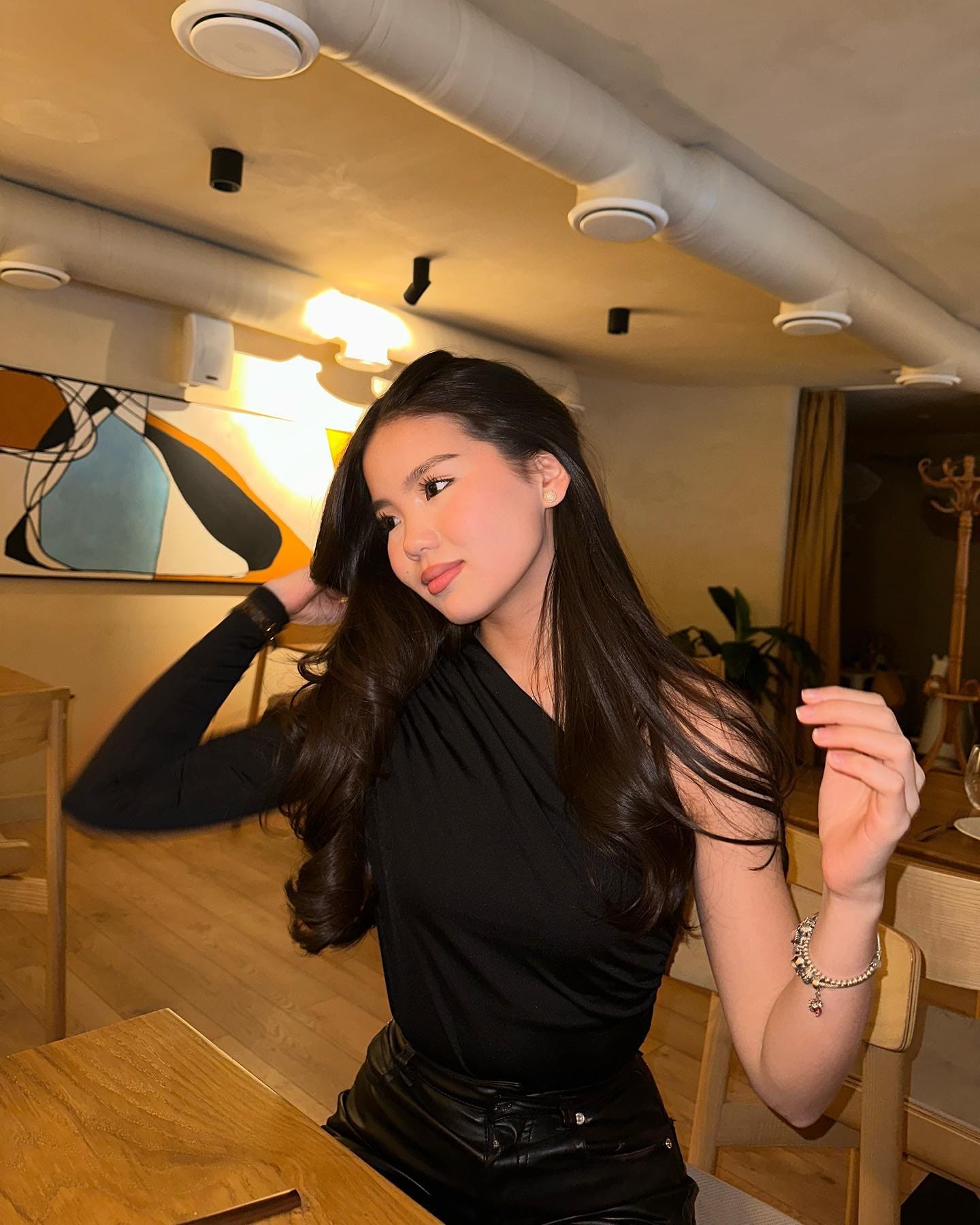 Dalina sits at a wooden table in a warmly lit room, wearing a black one-shoulder top and leather pants, with one hand near her hair.