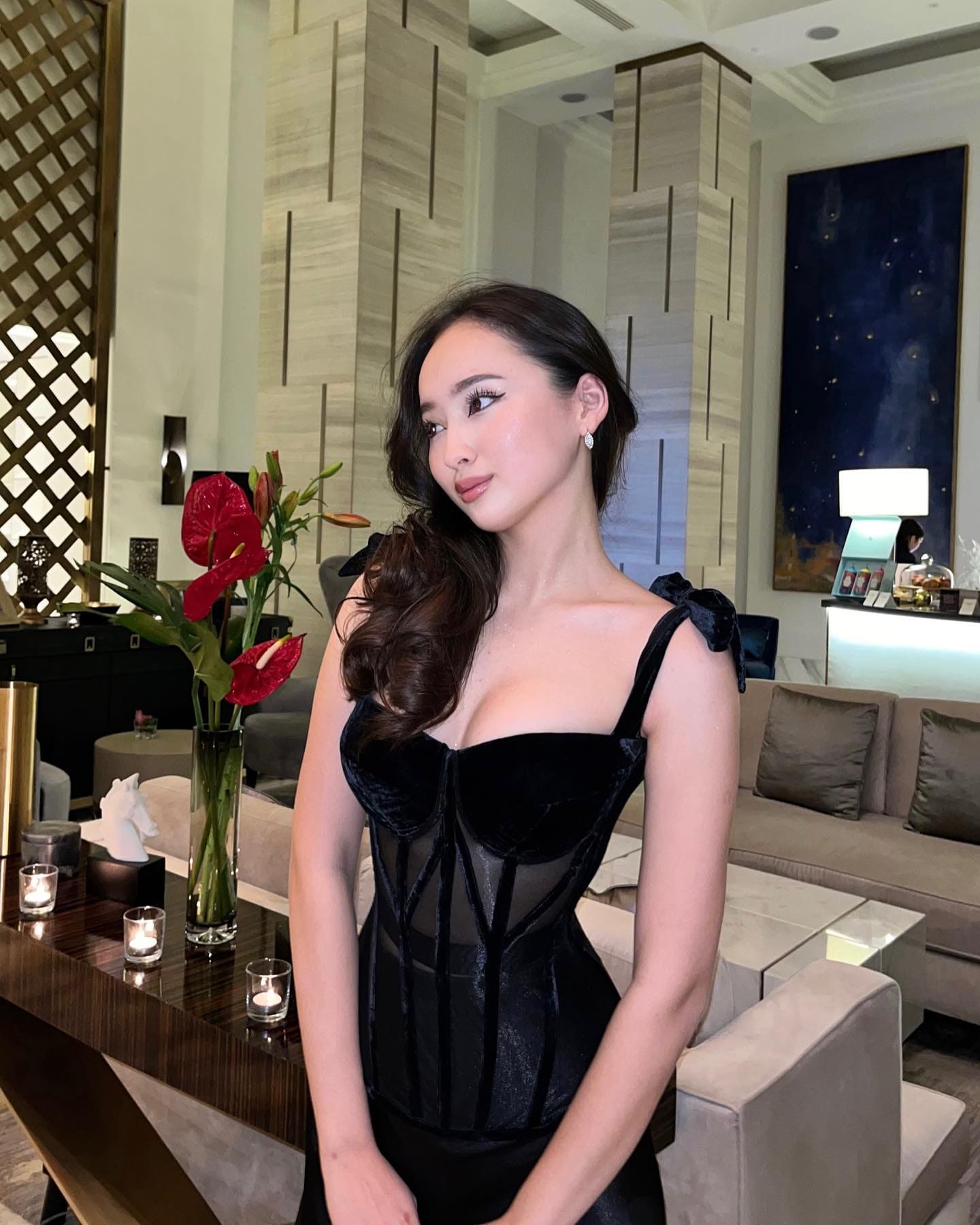 A woman in a black velvet dress with sheer panels poses in a modern lounge beside a table with red flowers and lit candles.