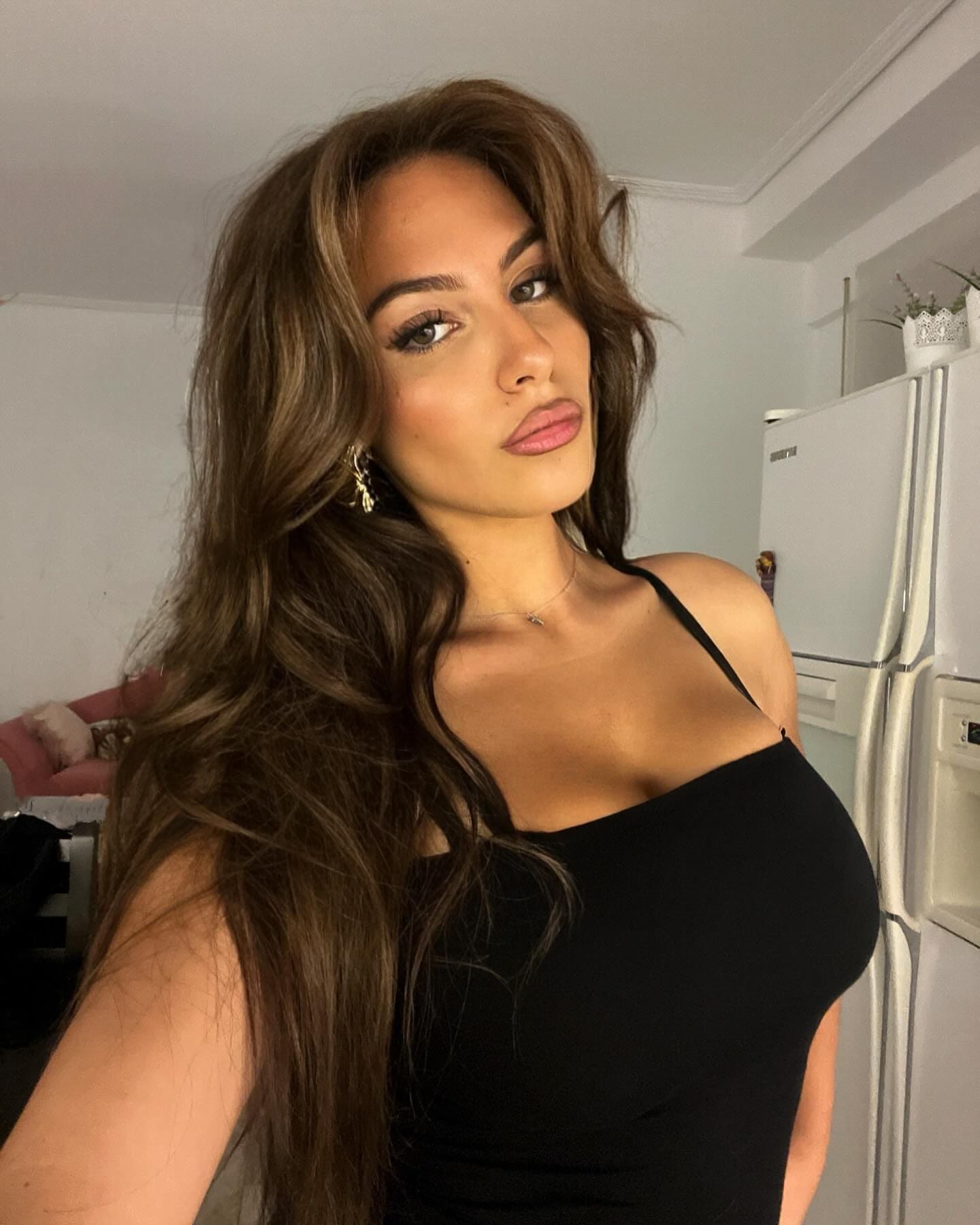 Loren in a black spaghetti-strap top, posing indoors near a white refrigerator and pink chair.