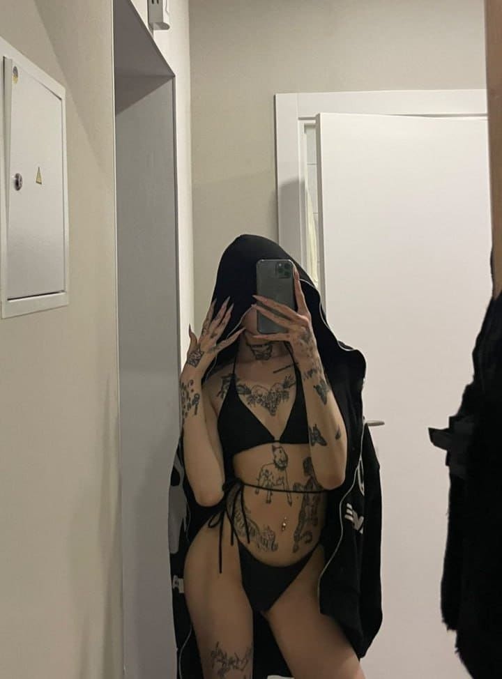 A tattooed person stands in a mirror selfie wearing a black string bikini and hooded jacket, holding a phone to cover their face in a white room.