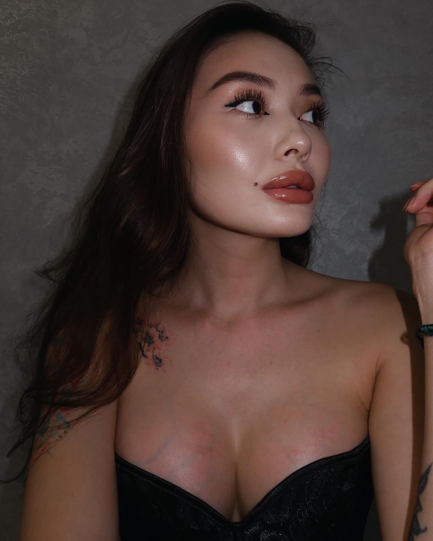 A woman with tattoos on her shoulder and arm wears a black strapless top with a deep neckline, looking to the side against a textured gray wall.