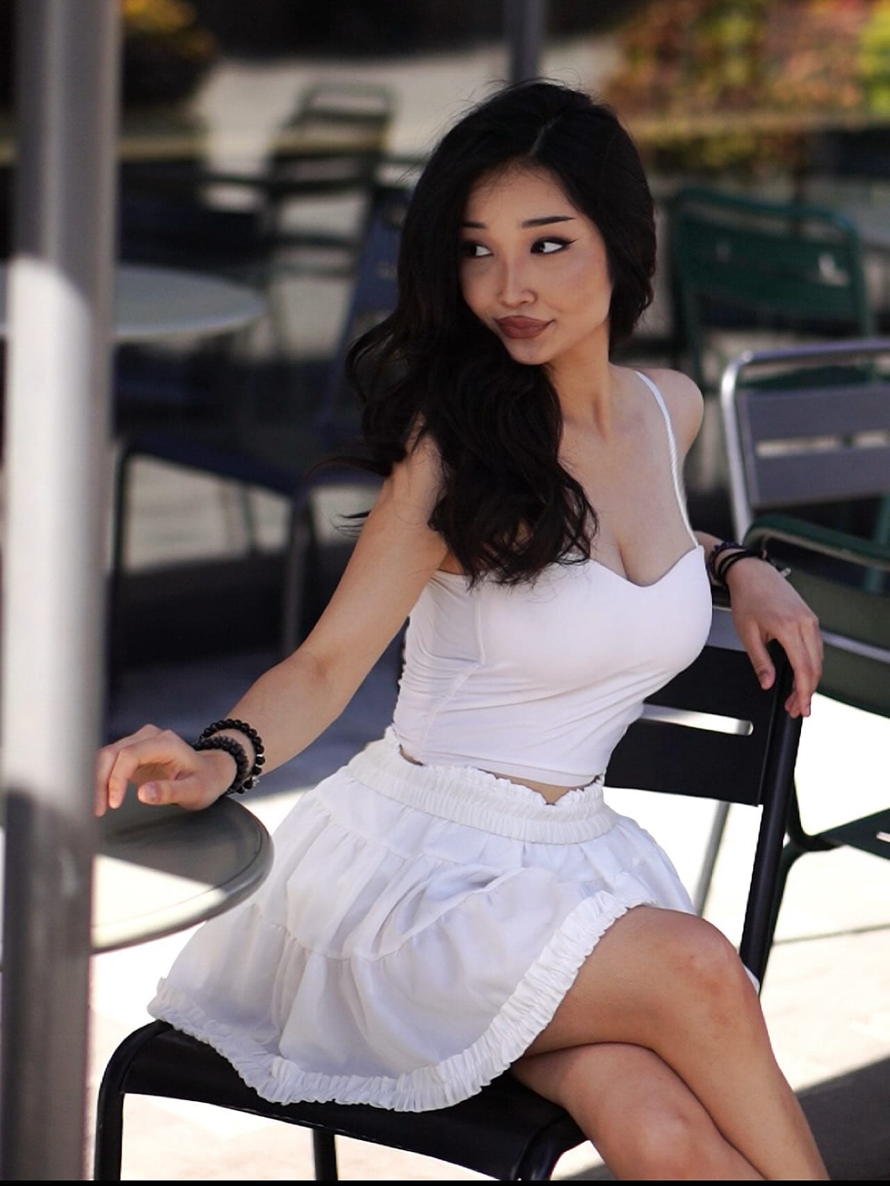 A woman with wavy hair sits on a black chair outdoors, wearing a white crop top and ruffled skirt.