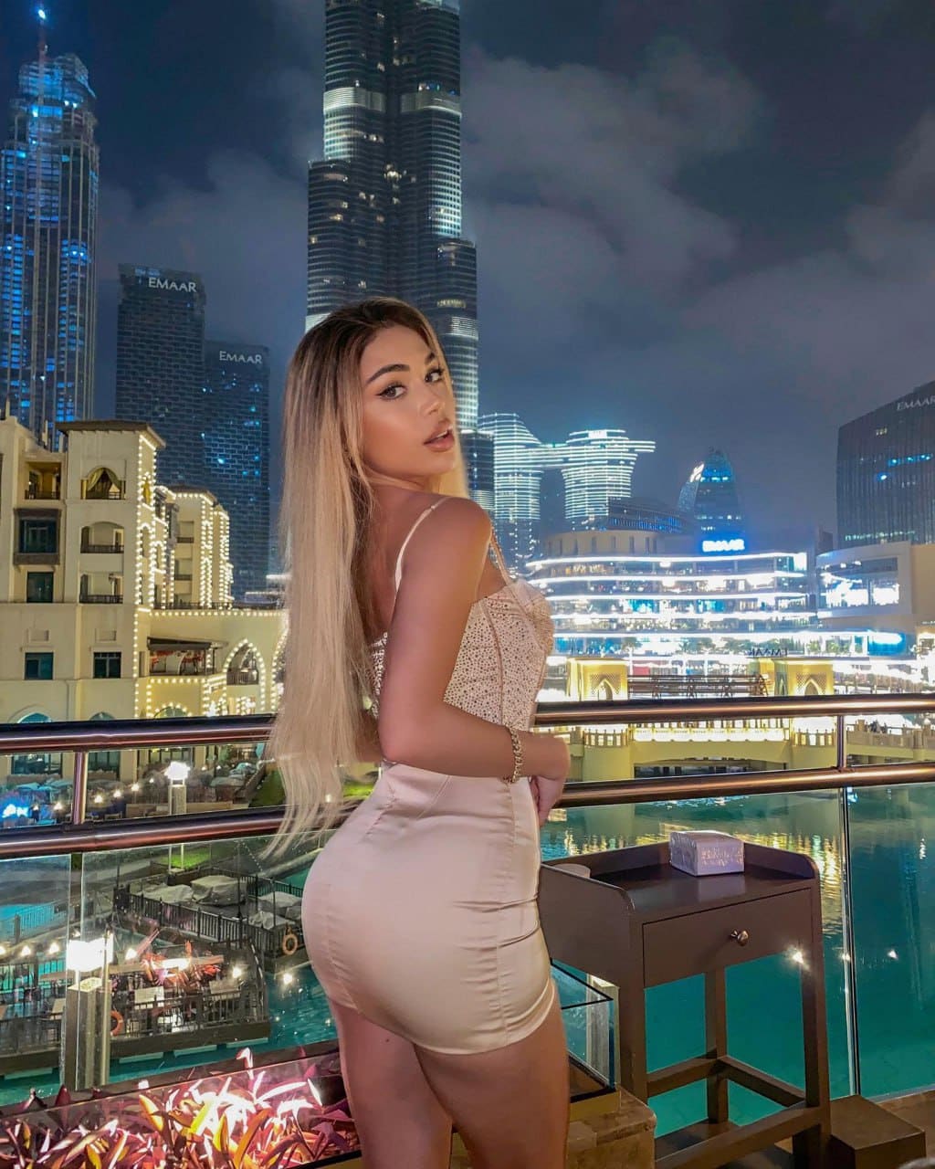 A woman stands in profile on a night rooftop terrace, wearing a short light-colored dress with thin straps against an illuminated skyline.