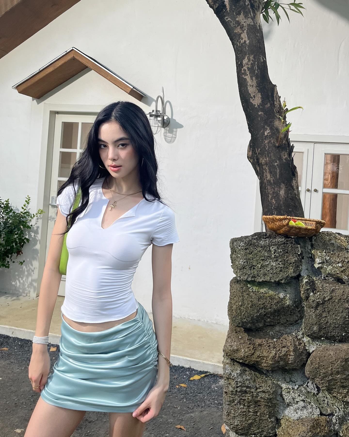 Standing by a white building, she wears a white top and light blue satin mini skirt, near a tree.