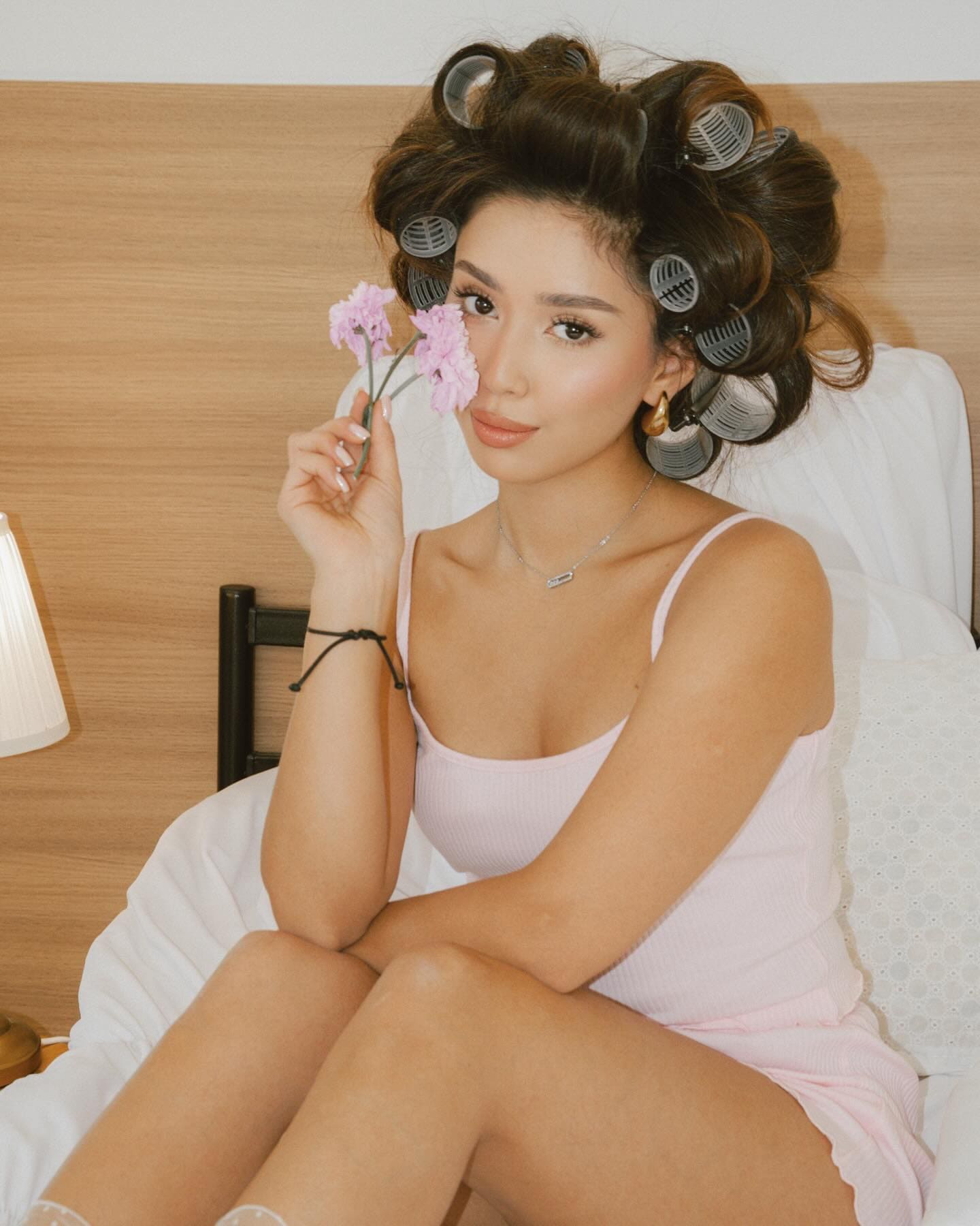 A woman with hair in curlers sits on a bed in a pink ribbed tank dress, holding two pink carnations near her face.