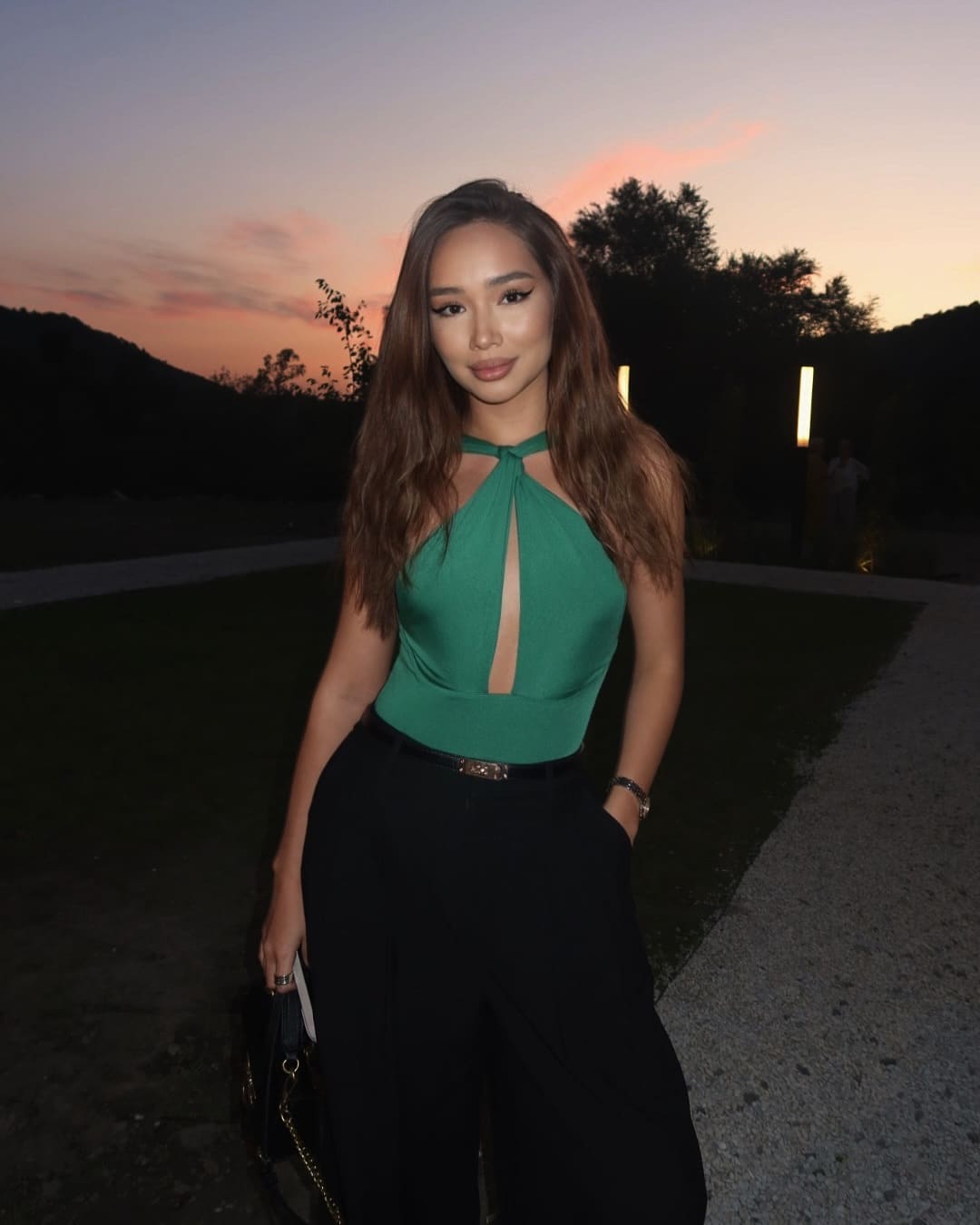 Woman in green halter top and black trousers stands at dusk, holding a black handbag with one hand in her pocket.