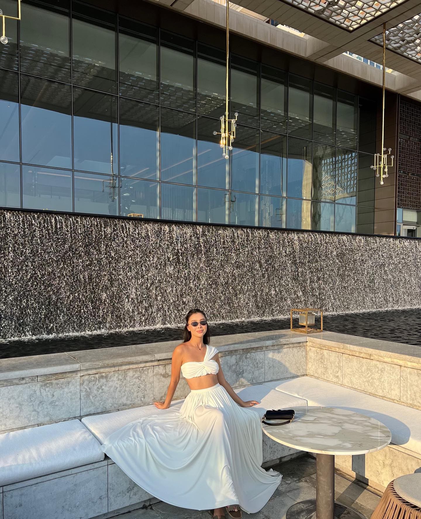 Woman in sunglasses, a white one-shoulder top, and long skirt sits on a bench near a water feature and glass wall.
