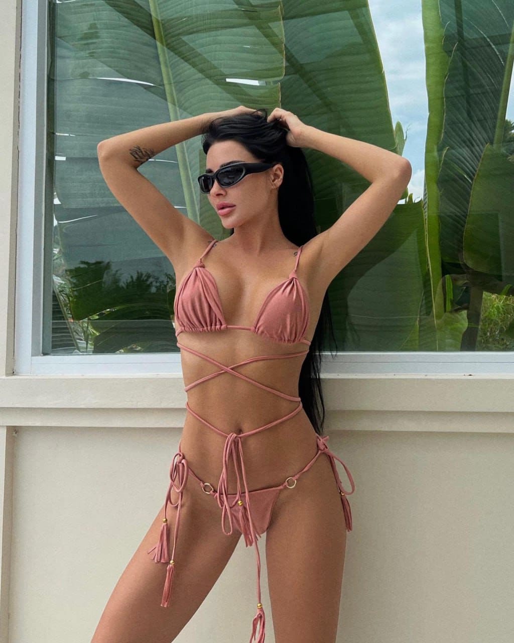A woman in a pink string bikini and sunglasses stands by a window with large green leaves, hands raised to her hair.