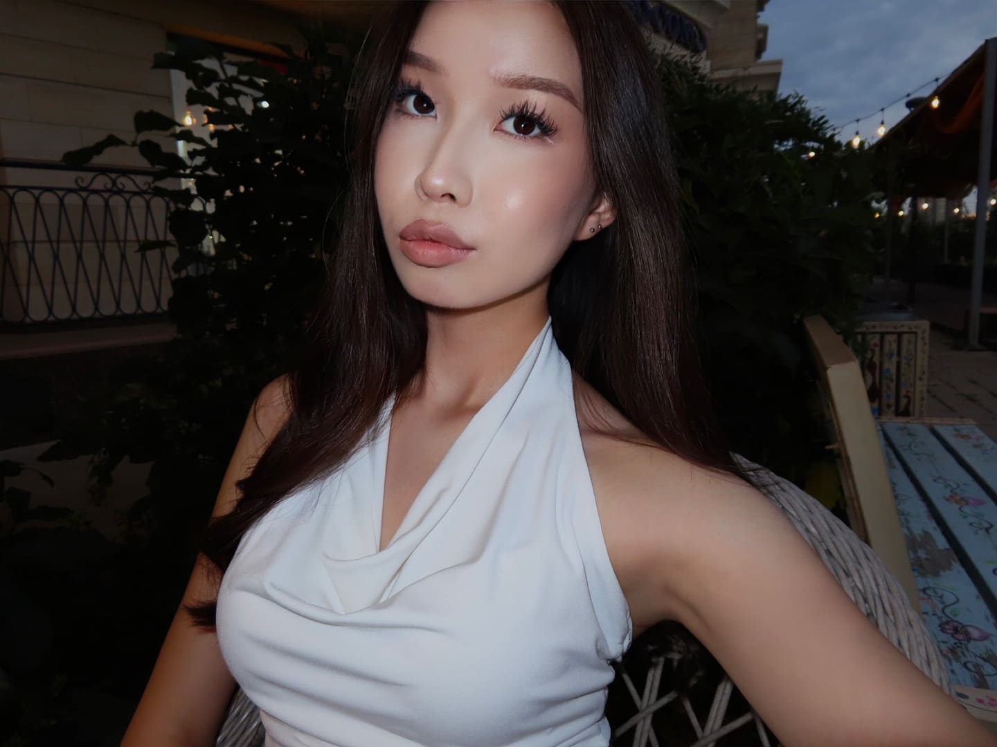 A woman sits outdoors at dusk in a white halter-neck top with a draped neckline, surrounded by greenery and string lights.