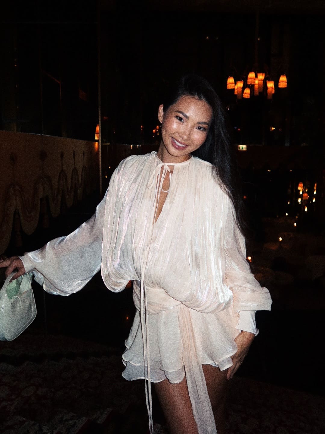 In a dim indoor space, she wears a loose sheer white blouse with a deep V-neck, holding a light-colored handbag.