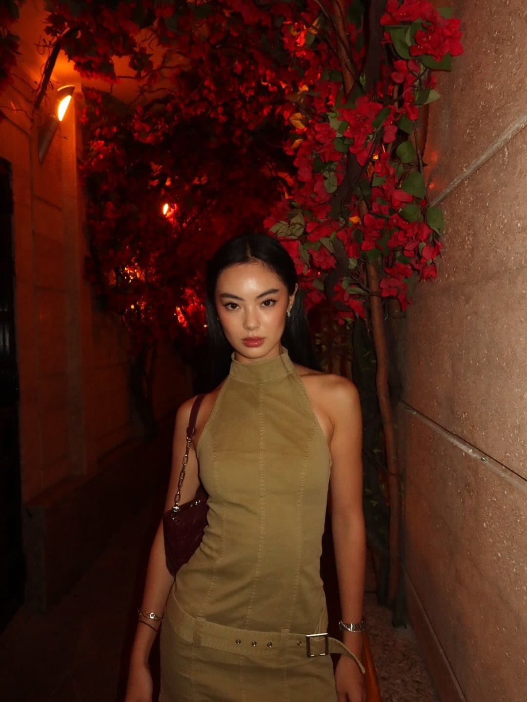 In a night passage, she wears an olive halter top and skirt with a belt, holding a bag near red vines.