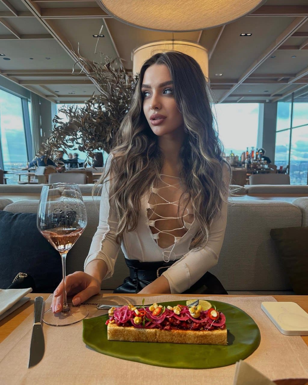 A woman with wavy brown hair sits at a restaurant table in a white lace-up top, holding a glass of rosé wine.