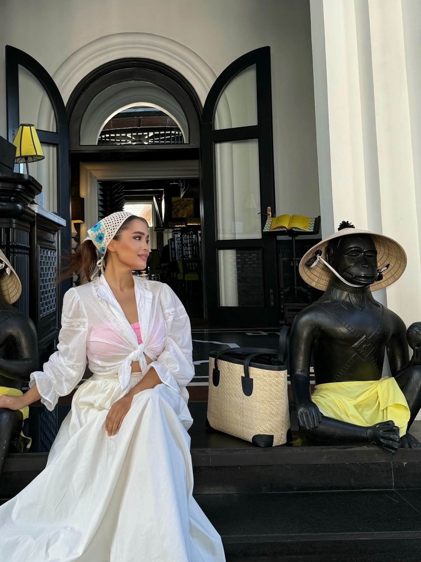 A woman in a white blouse, pink top, and long skirt sits on a stone step next to a black statue, before an arched doorway with black frames.