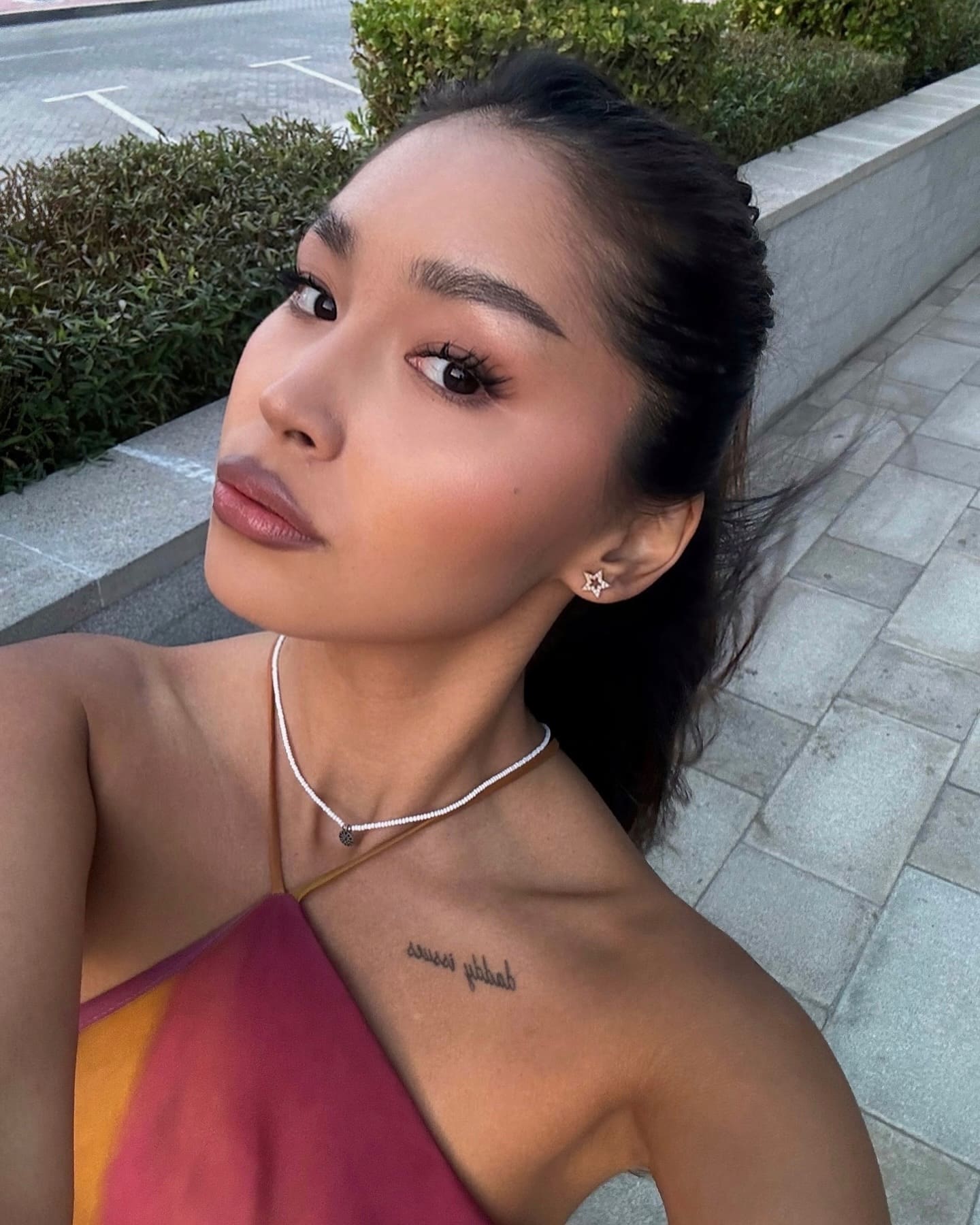A woman with a ponytail and star earring wears a multicolored top and necklace on a paved walkway.