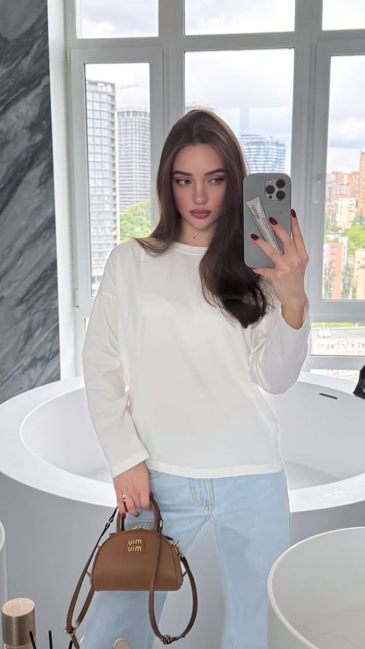 A mirror selfie in a bathroom shows a woman in a white long-sleeved top and light blue jeans, holding a brown handbag near a bathtub.