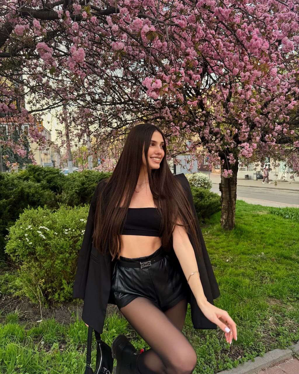 Liana stands outdoors by a pink tree in a black crop top, leather shorts, tights, and blazer, with one hand near her knee.