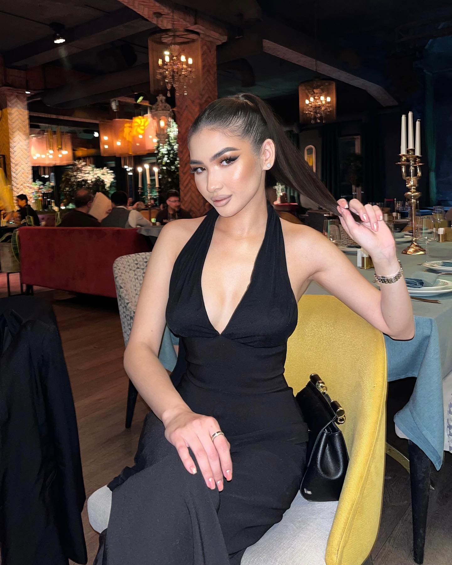 Meilin sits in a restaurant in a black halter-neck dress with a deep V neckline, one hand raised to her hair and a black handbag beside her.