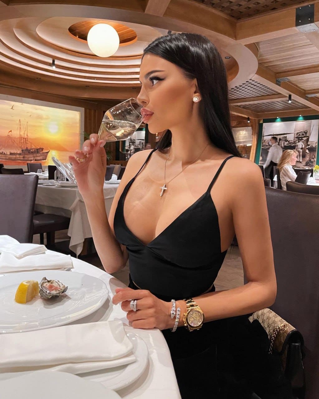 A woman in a black spaghetti-strap top holds a glass of water at a restaurant table, with an oyster and lemon wedge on a plate.