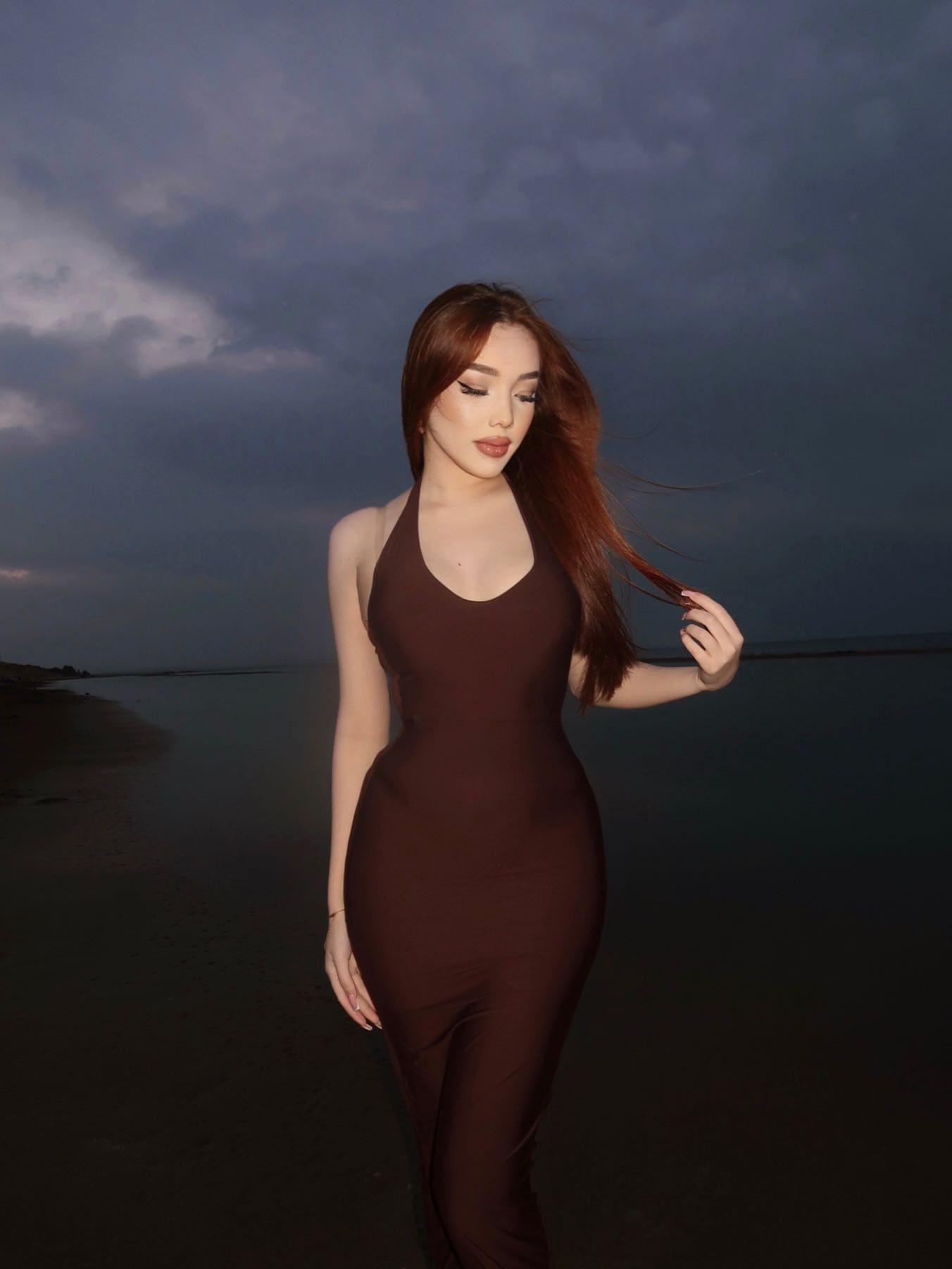 A woman stands on a beach at dusk in a dark brown halter dress, holding a strand of hair and looking down against the evening light.