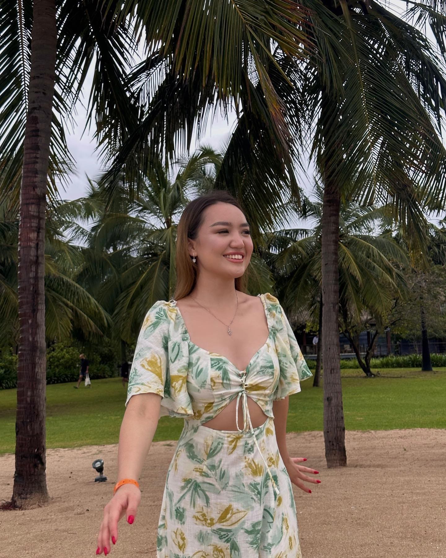 A woman with long brown hair smiles on a sandy path before palm trees, wearing a floral-print two-piece outfit with a midriff cutout.