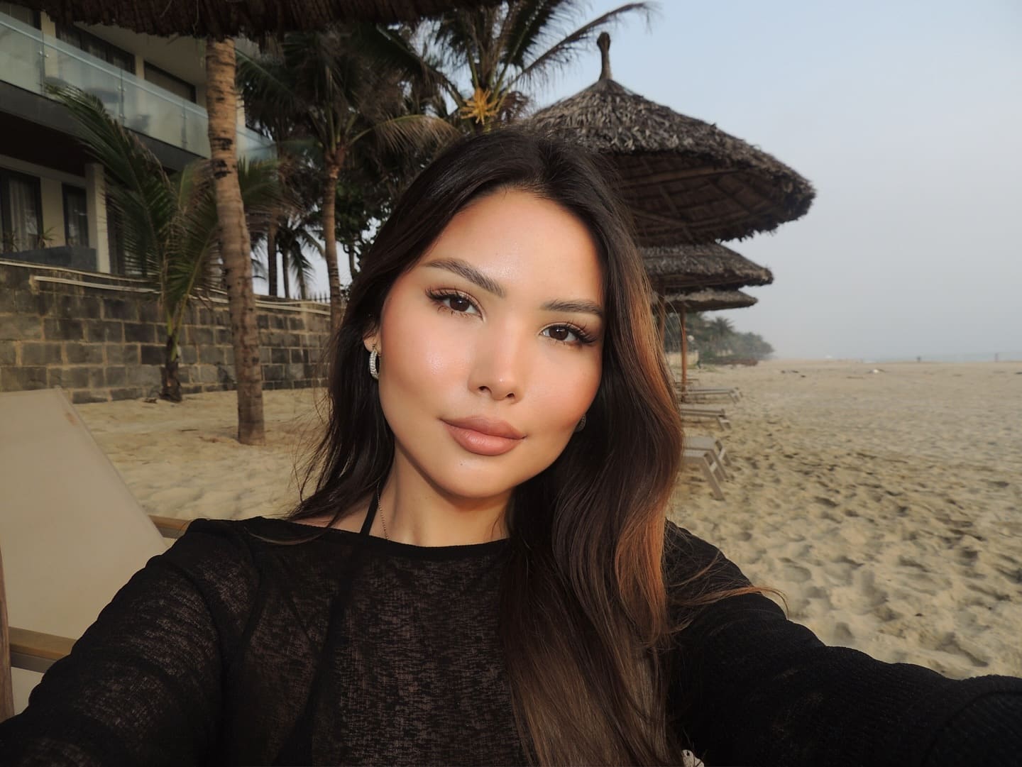A selfie on a sandy beach shows a woman in a sheer black top, with palm trees and umbrellas in the background.