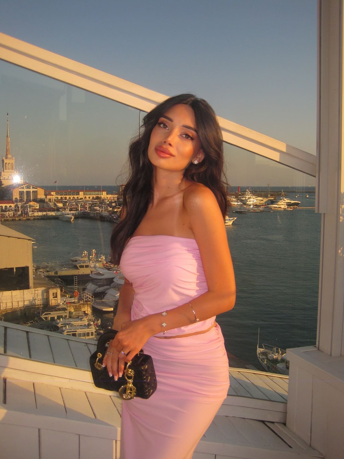 A woman in a pink two-piece outfit stands on a balcony holding a black handbag, with boats visible in the sunset background.