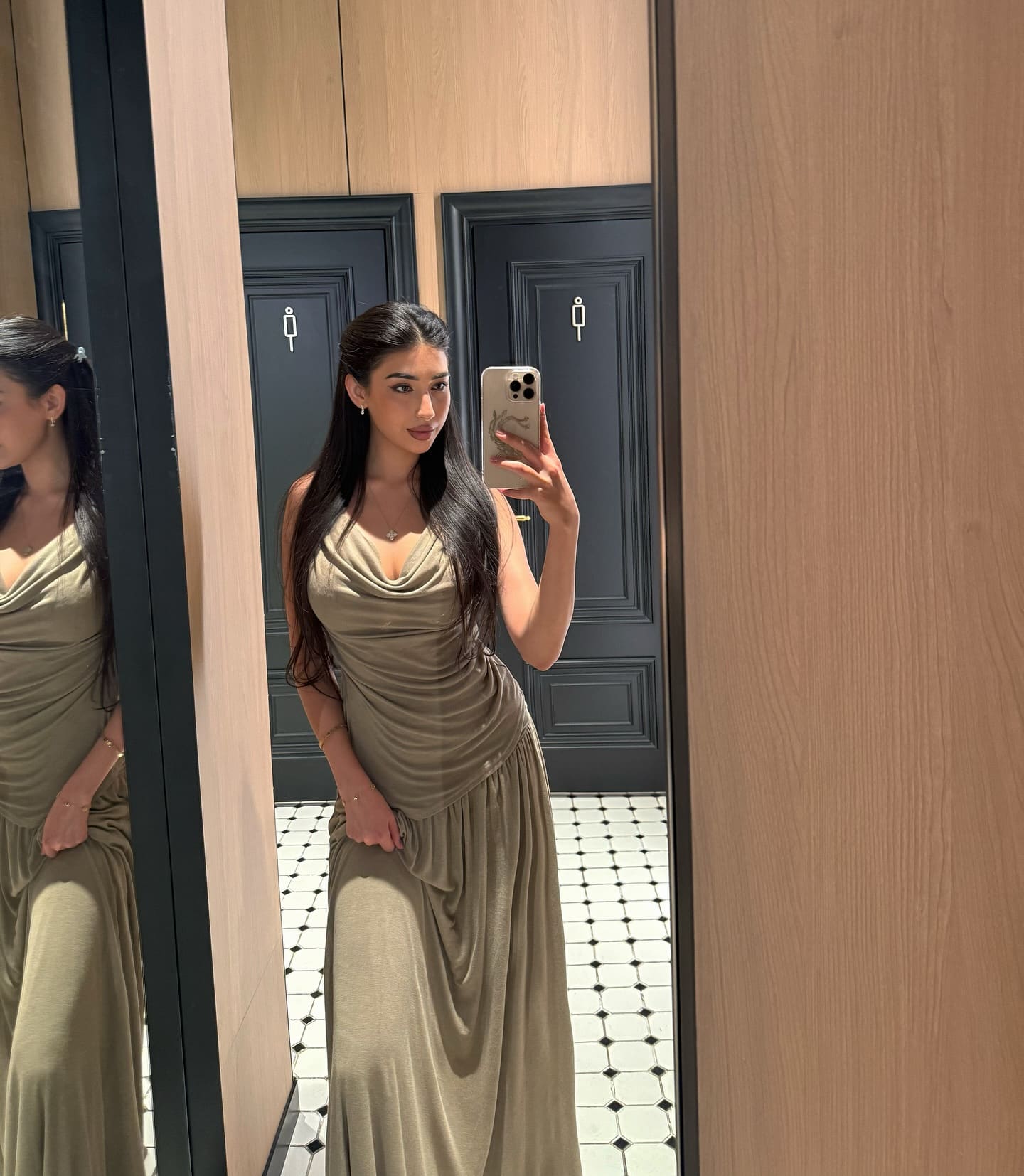 A mirror selfie captures a woman in an olive-green draped maxi dress, standing in a room with wood paneling and tiled floors.