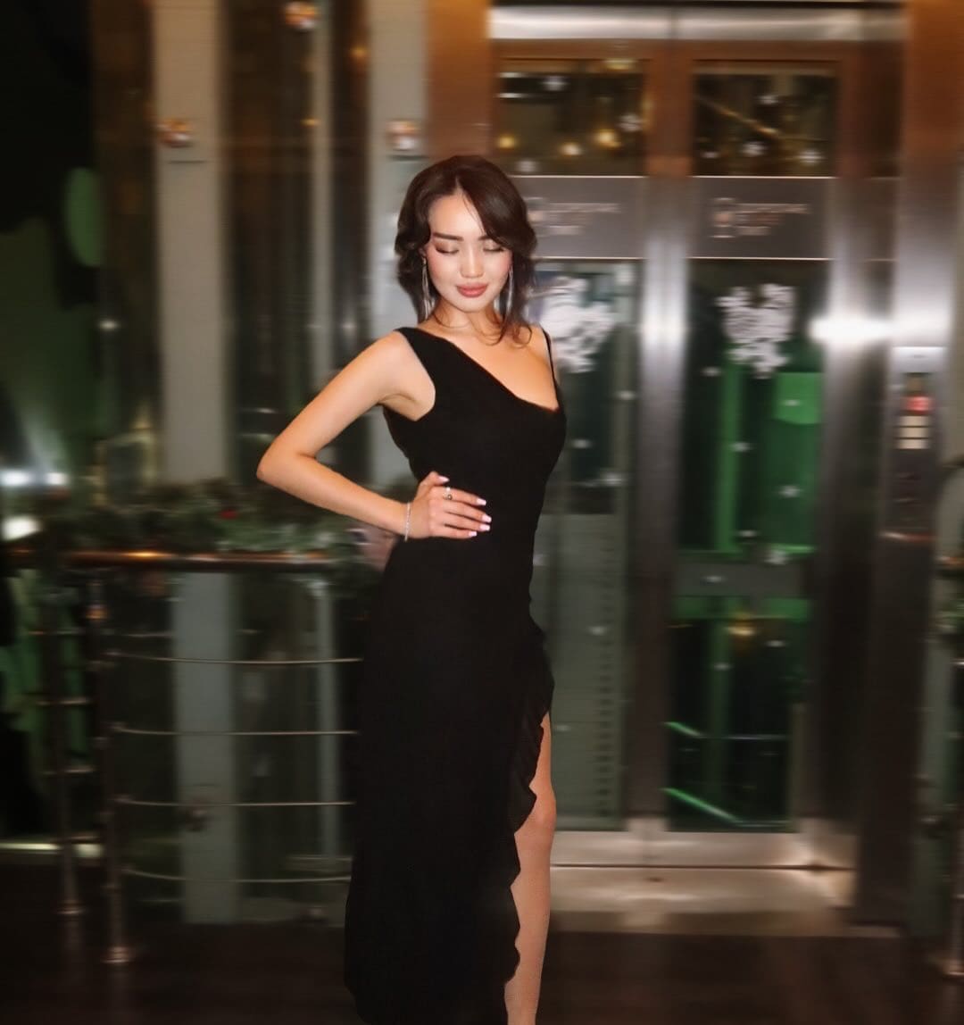 A woman in a sleeveless black floor-length dress with a high side slit stands in an indoor lobby, hand on hip, head tilted down.