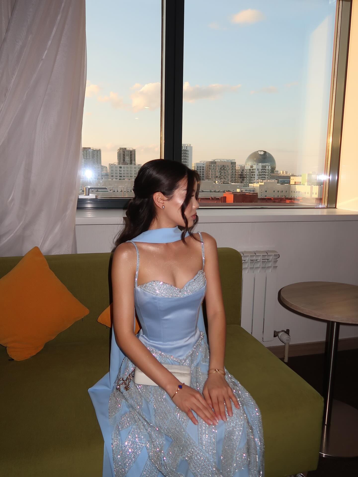 A woman in profile sits on a green sofa in a light blue satin gown with a sequined skirt, with a city skyline visible at dusk.