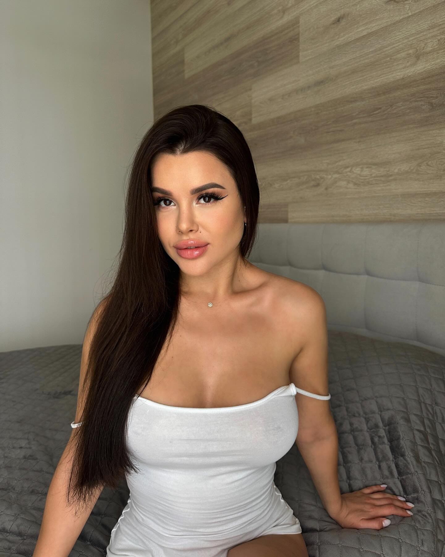 Tina sits on a gray quilted bed in a white strapless mini dress, with a wood-paneled wall behind her.