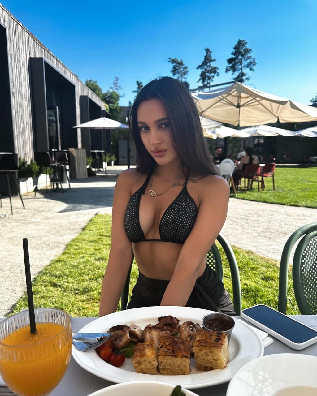 Tatiana sits at an outdoor table in a black patterned bikini, with food and orange juice under a clear blue sky.