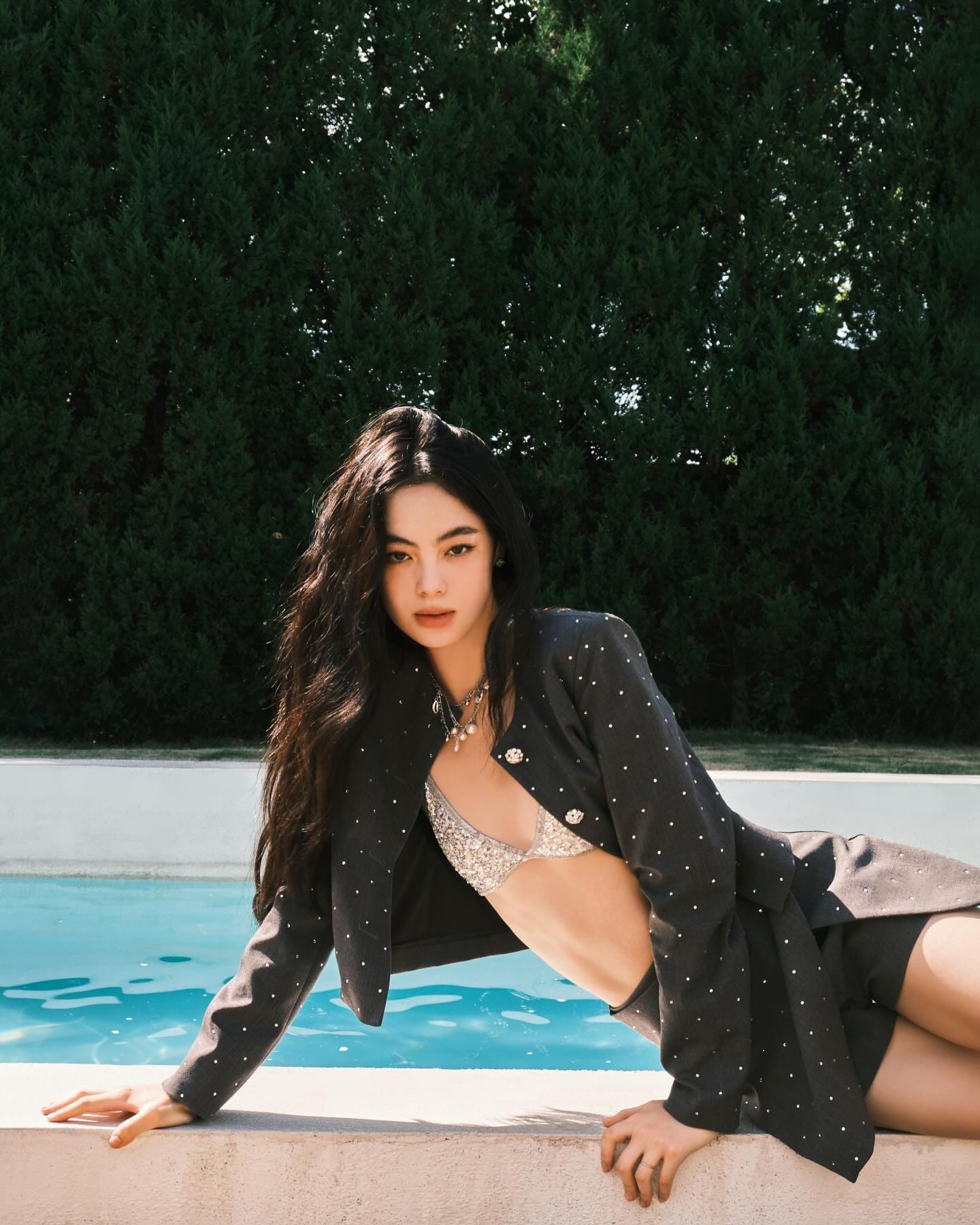 She leans on a pool edge in a black studded jacket and silver bra top, with a green hedge behind her.