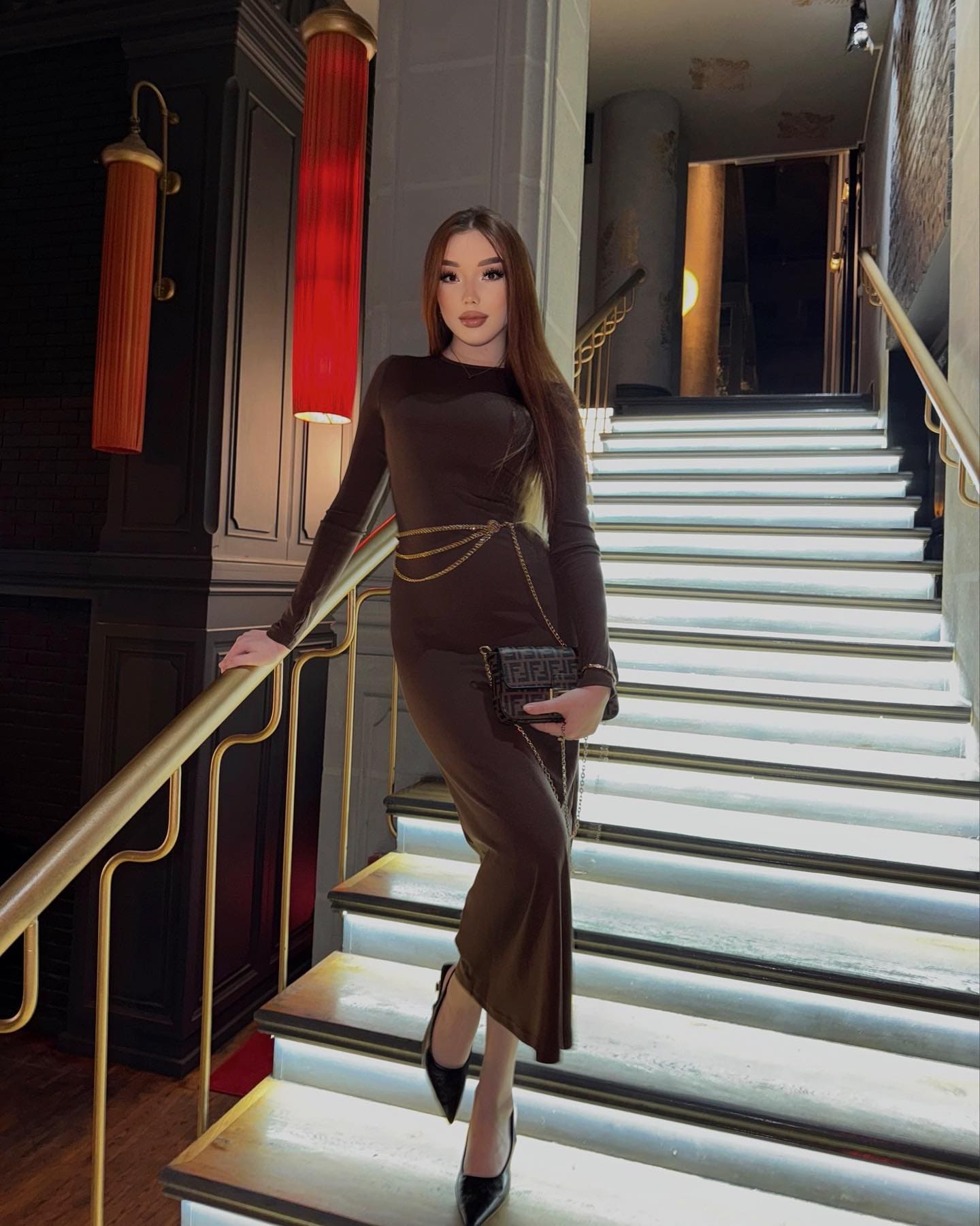 A woman stands on an illuminated staircase in a brown midi dress with a gold belt, holding a clutch and resting a hand on a brass railing.