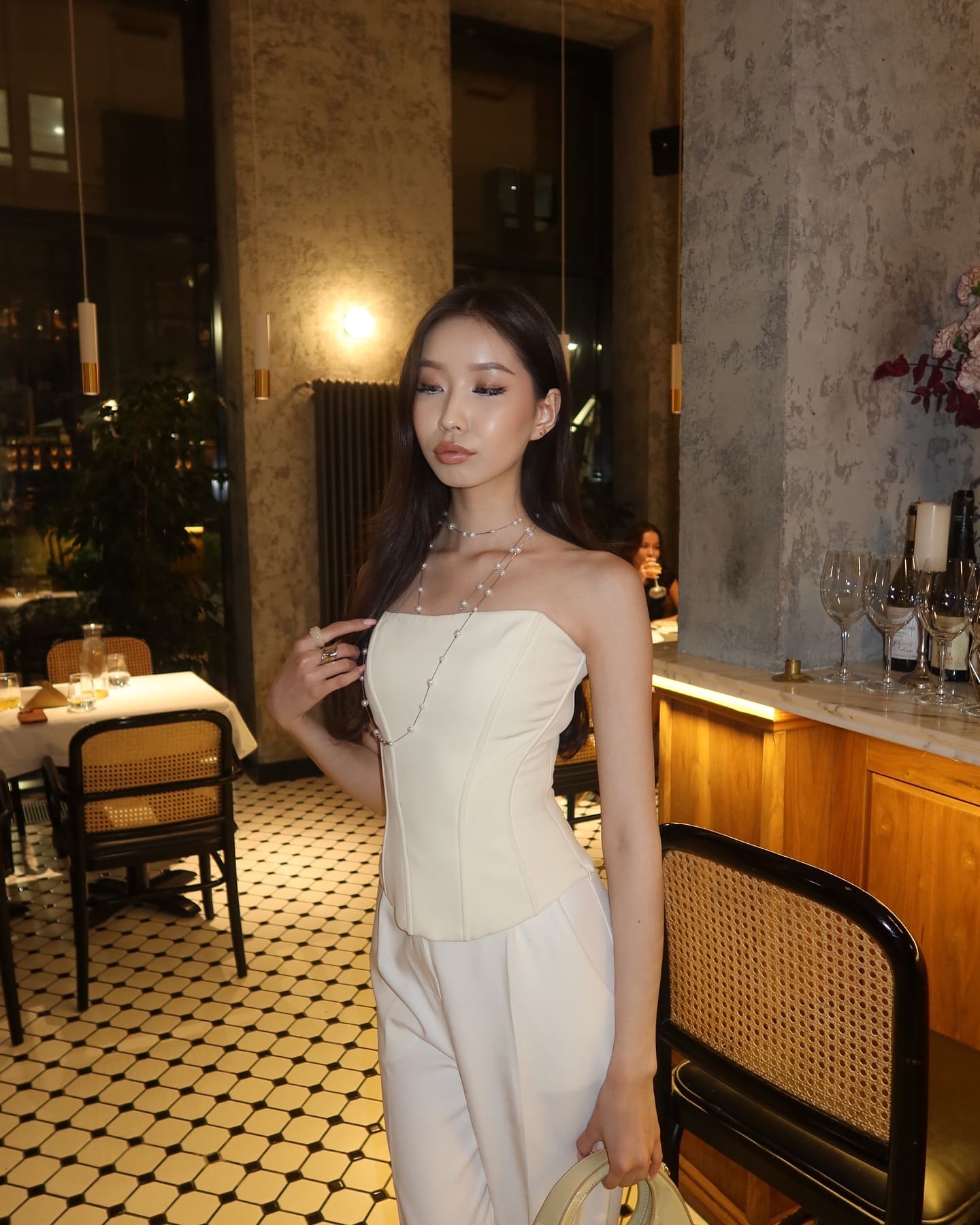 Lysandra stands in a restaurant wearing a white strapless top, white trousers, and a long beaded necklace, holding a light handbag.
