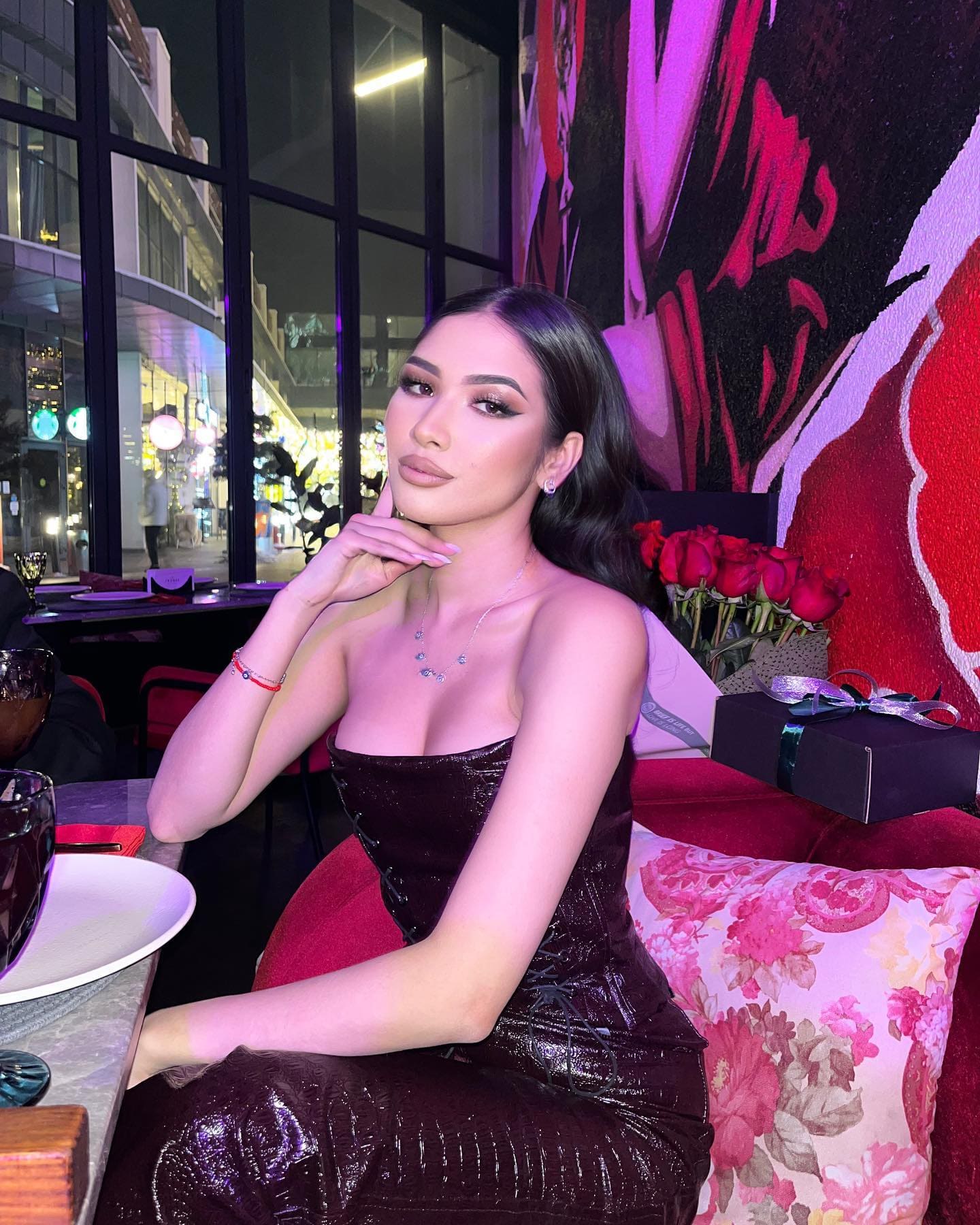 A woman in a strapless dark sequined dress rests her chin on her hand at a table, with red roses and a city view at night behind her.