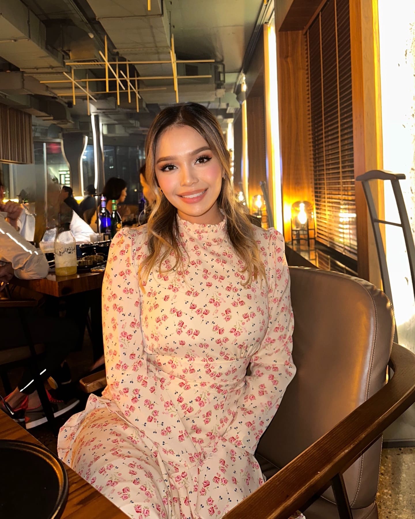 A woman with wavy hair smiles in a brown leather chair, wearing a cream floral dress, inside a warmly lit restaurant with wooden paneling.