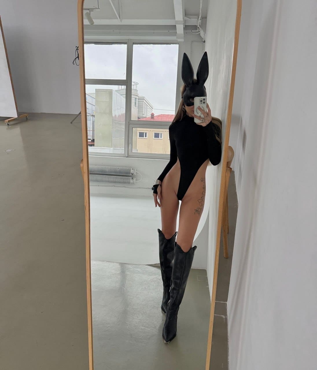 A person in a black bunny mask, bodysuit, and knee-high boots takes a mirror selfie in a room with a large window.