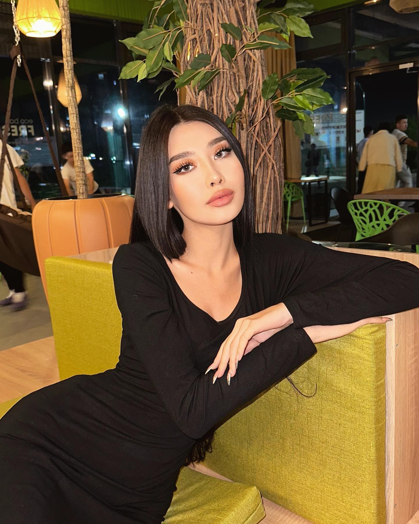 Seated on the same green bench, she wears a black V-neck dress and leans on the backrest, looking directly at the camera.