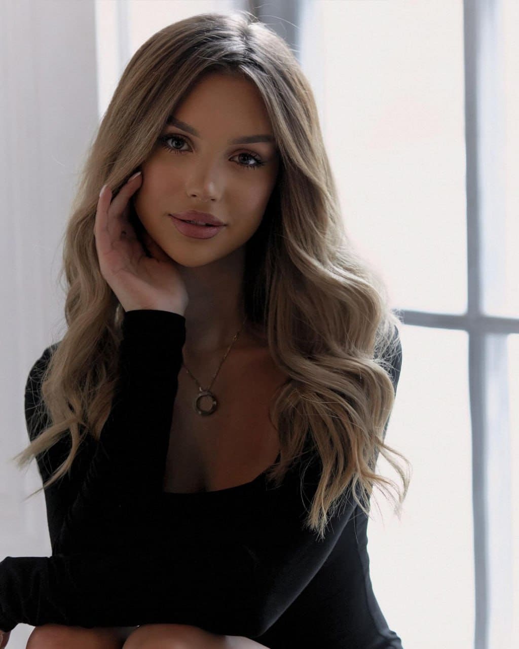 A blonde woman sits indoors near a window, wearing a black long-sleeved top with a deep neckline and a pendant necklace.