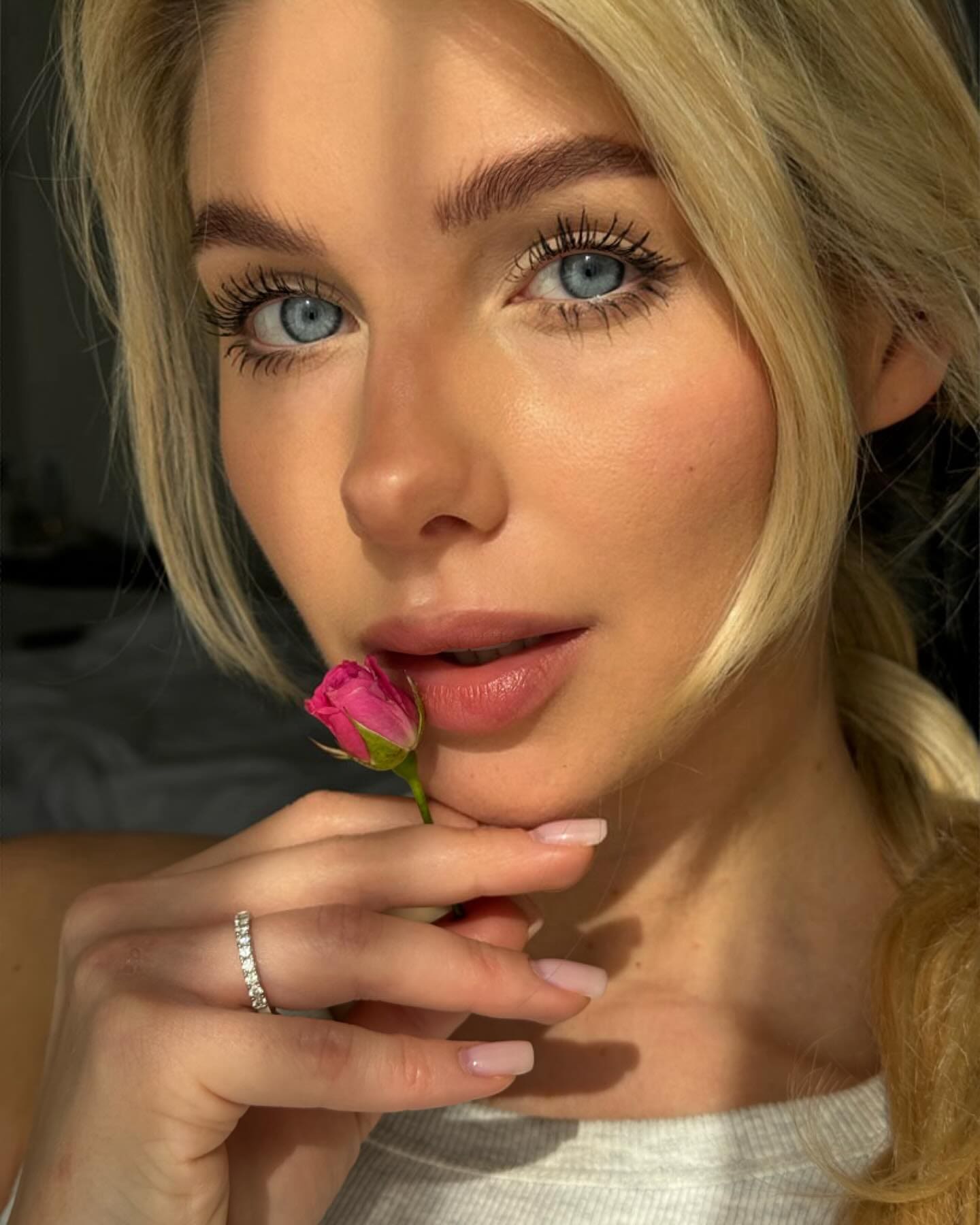 Close-up of a blonde woman holding a pink rosebud to her lips, wearing a white ribbed top and diamond ring.