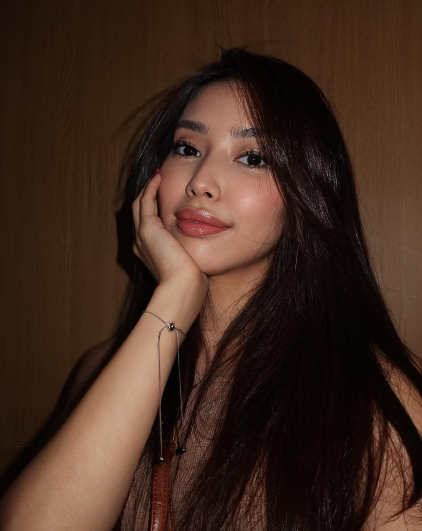 A woman rests her chin on her hand, wearing a sheer sleeveless top and thin bracelet, against a plain brown background.