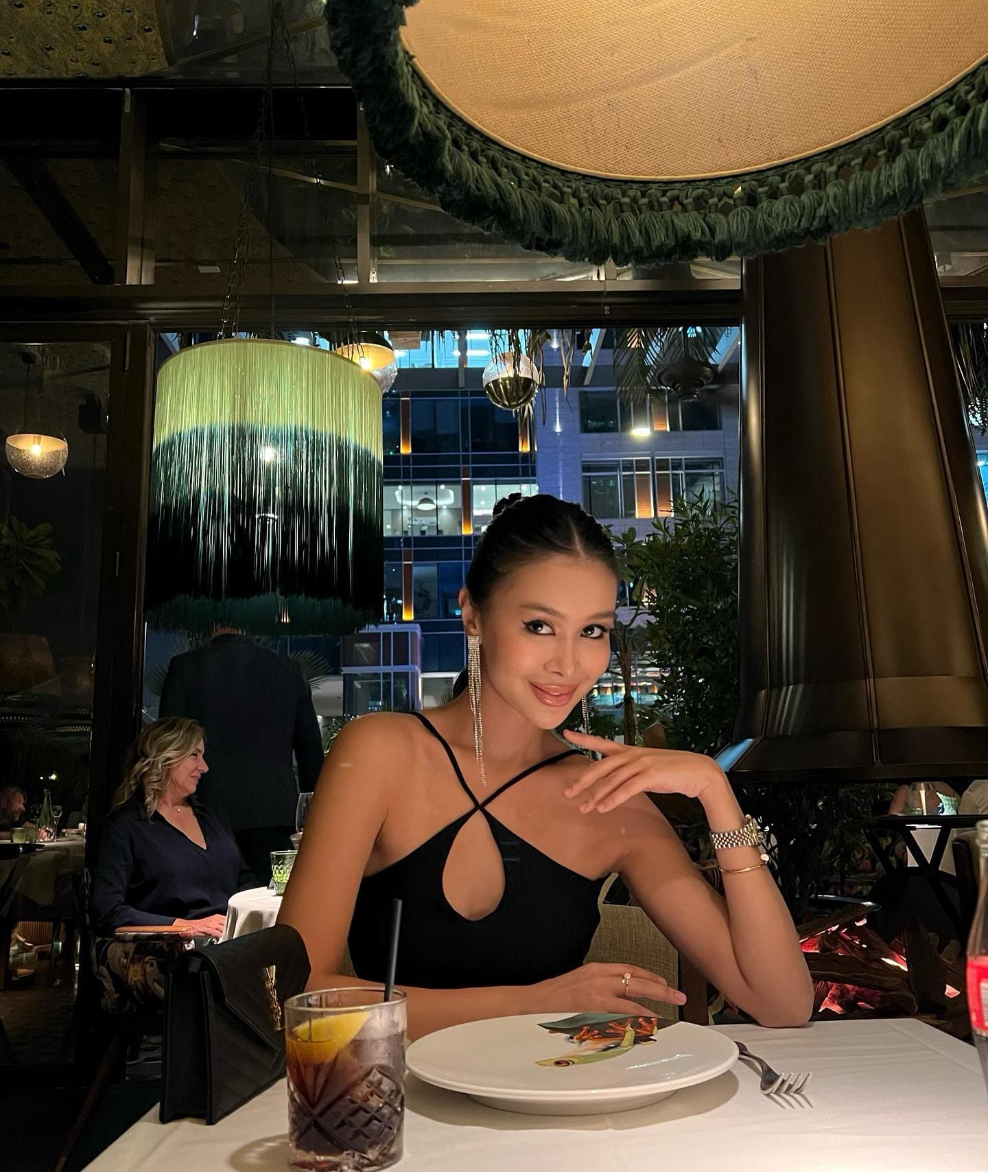 Woman in a black strappy keyhole top and earrings sits at a restaurant table with food and a glass in front of her.