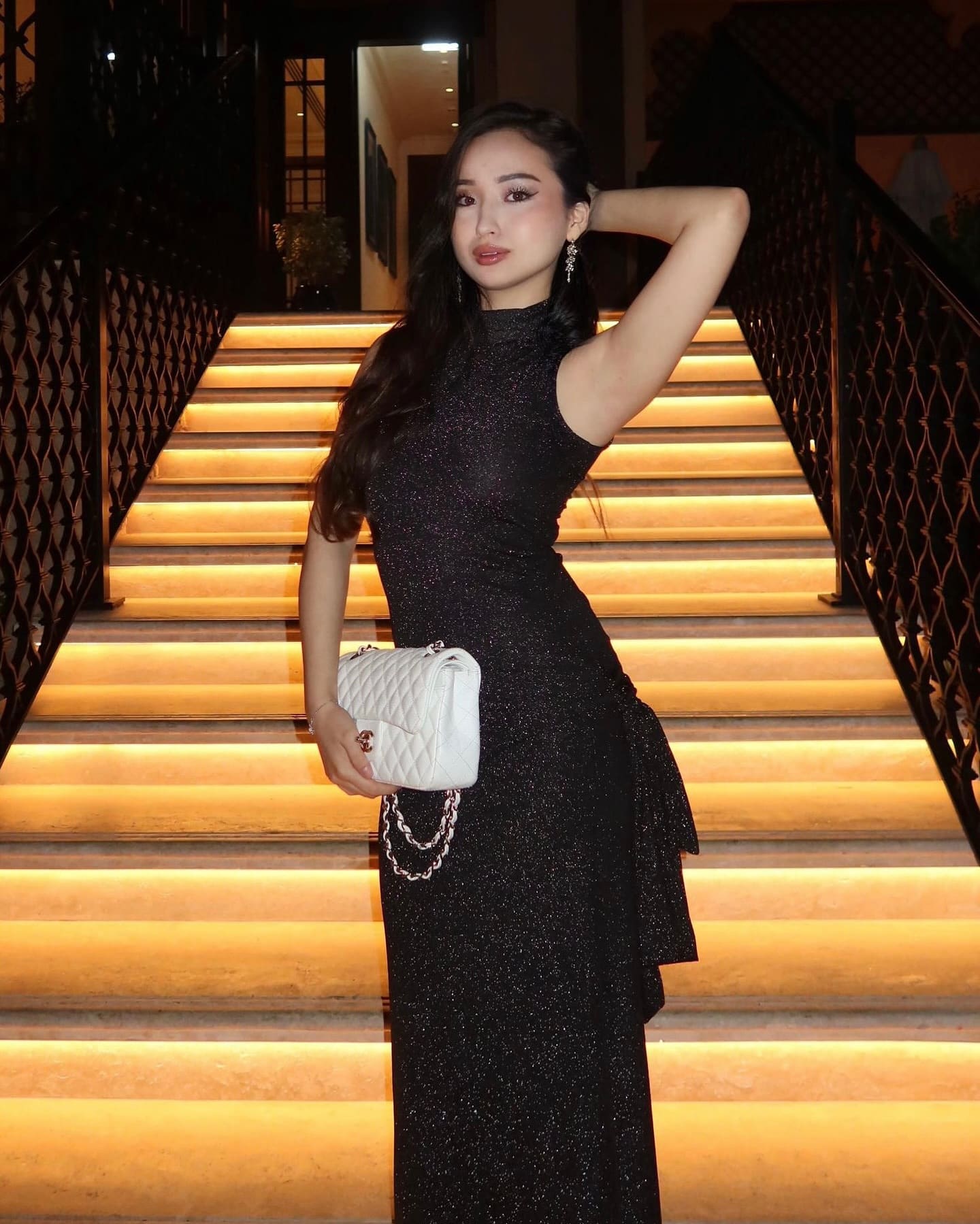 A woman in a sparkly black high-neck gown stands on a lit staircase, holding a white bag with one arm raised behind her head.