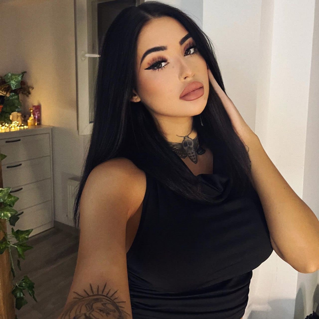 Amira in a black sleeveless top poses indoors with a hand in her hair, showing arm and neck tattoos near a white dresser.