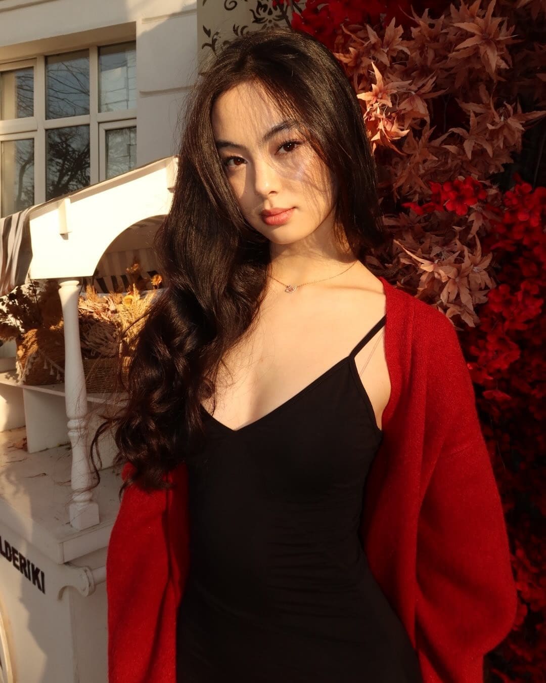 Ilaria stands outdoors in a black top and red cardigan, with a necklace, against white structures and red foliage.