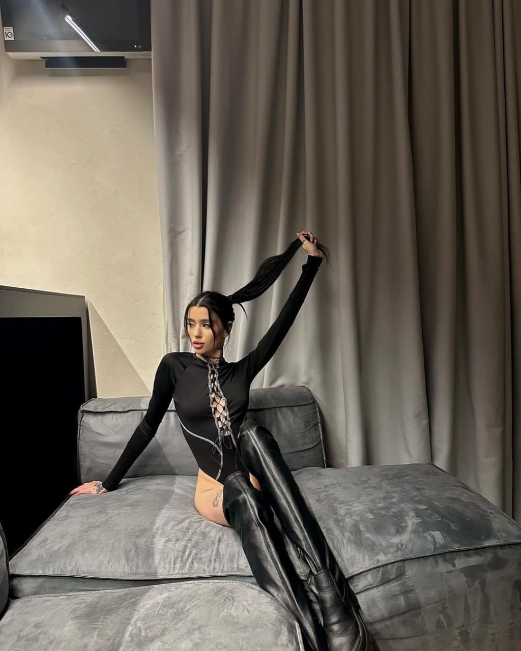 A woman with long dark hair sits on a grey sofa in beige curtains, wearing a black lace-up top and thigh-high boots, holding her hair up.